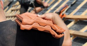 Ein Mann trägt Dachpfannen. Im hintergrund ist ein ungedecktes Dach zu sehen