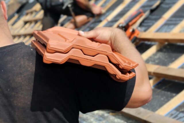 Dachdecker Ein Mann trägt Dachpfannen. Im hintergrund ist ein ungedecktes Dach zu sehen